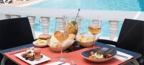 Mesa de comedor con diversos platos y bebidas, con vista a una piscina.