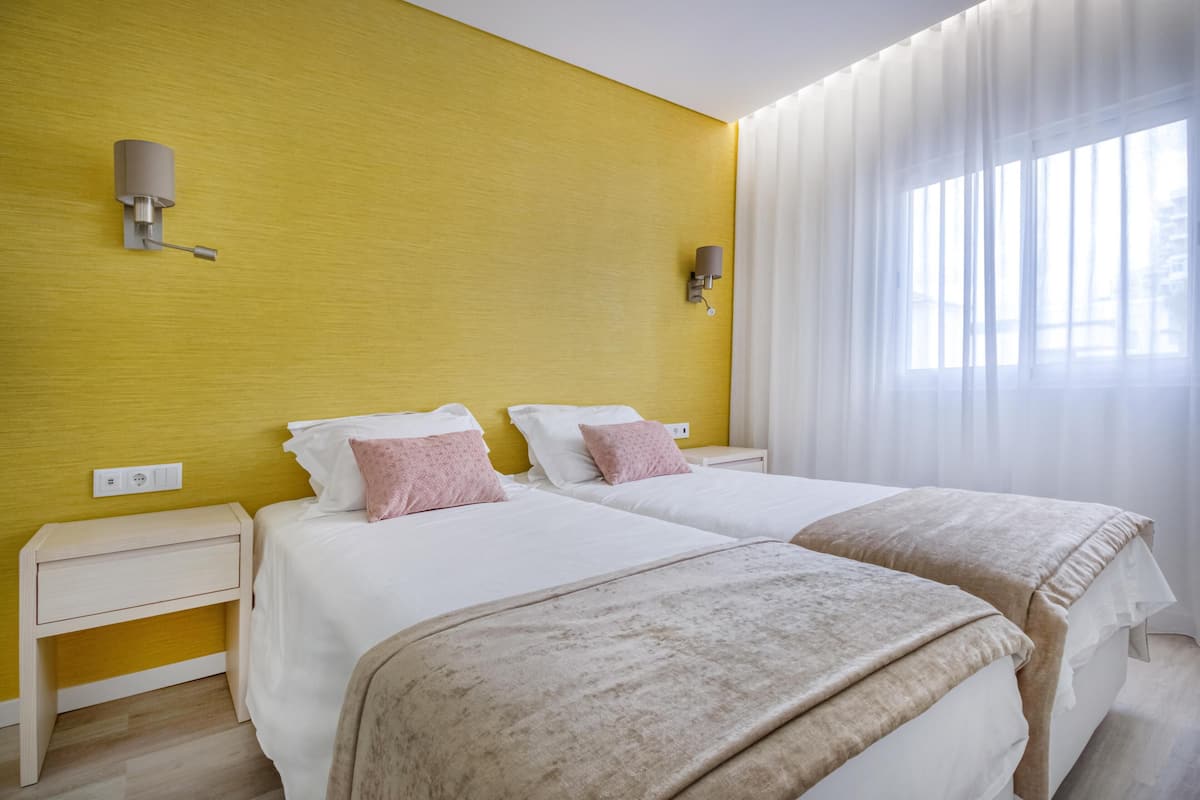 Habitación de hotel con dos camas, una pared de acento amarilla y un ventanal con cortinas traslúcidas.