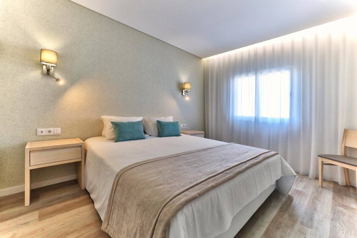 Una habitación de hotel moderna con una cama grande, dos mesitas de noche y una mesita auxiliar con cajón. Hay dos lámparas de pared y una ventana con cortinas.