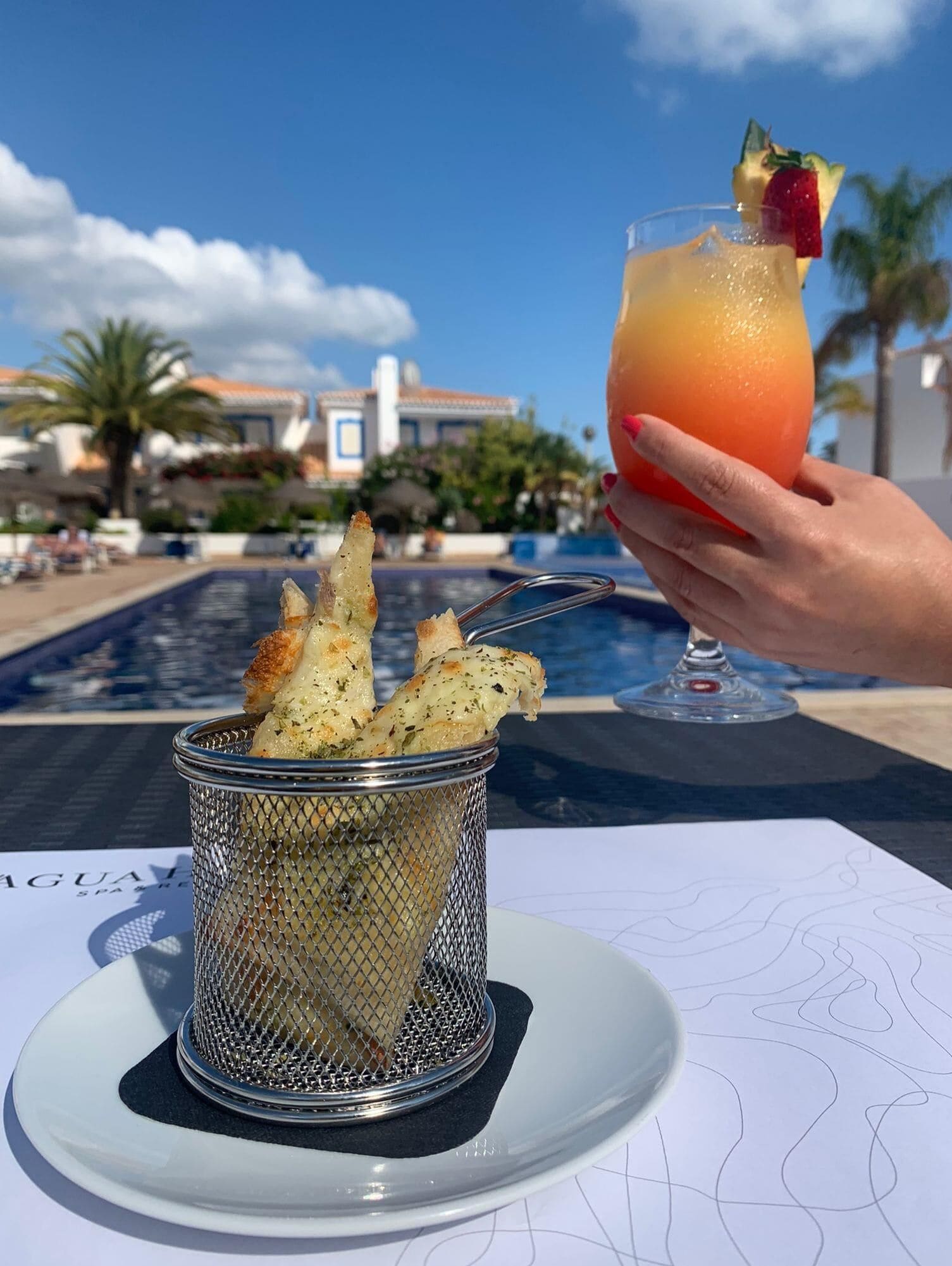 A hand holding a cocktail with a garnish, a basket of garlic bread, and a poolside view.