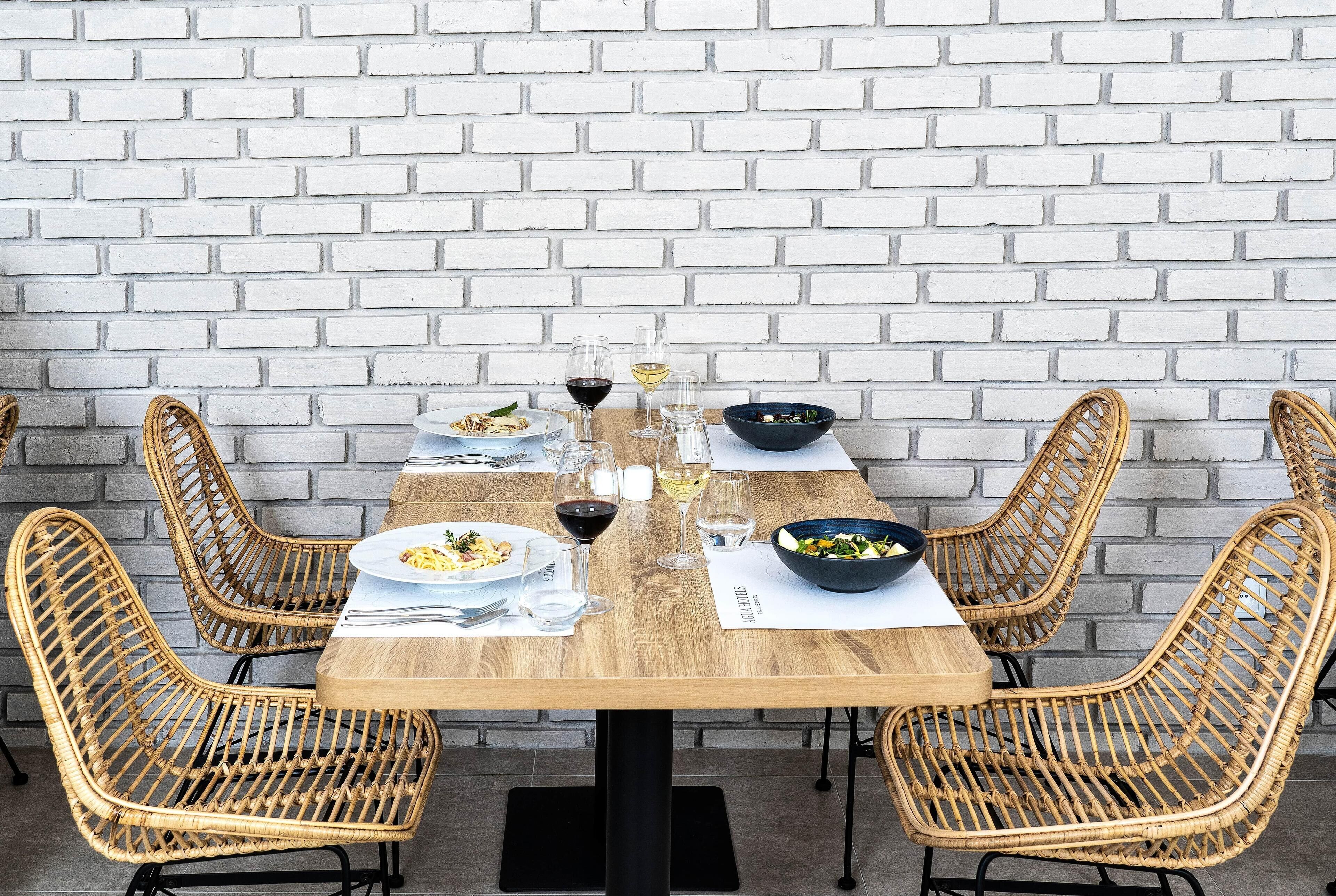 Una mesa de comedor con platos, vasos y un tazón de ensalada, rodeada de sillas de mimbre, frente a una pared de ladrillo blanco.