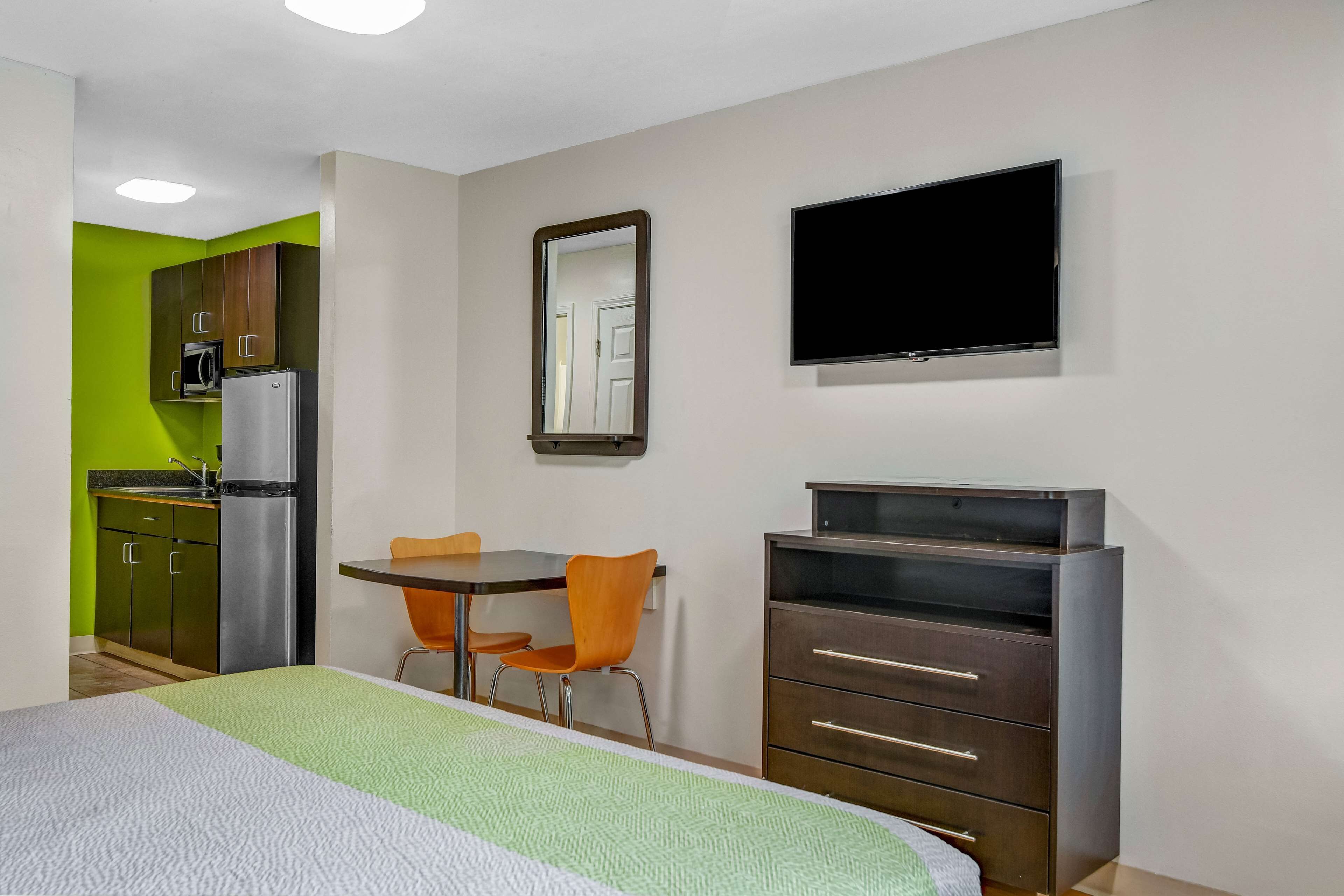 A hotel room with a bed, a desk, a chair, a TV, a dresser, and a kitchenette with a fridge and microwave.