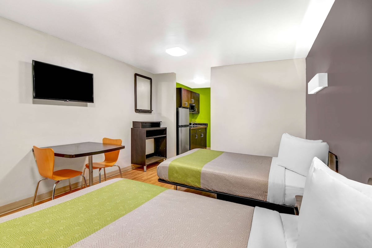 A hotel room with two beds, a desk, chairs, a TV, and a kitchenette.