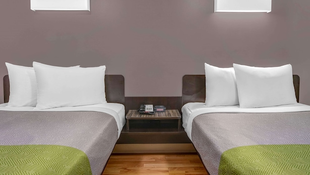A hotel room with two beds, a small bedside table with a phone and a clock, and a wall-mounted light fixture. - Studio 6 Extended Stay - Atlanta, GA - Chamblee (Chamblee)