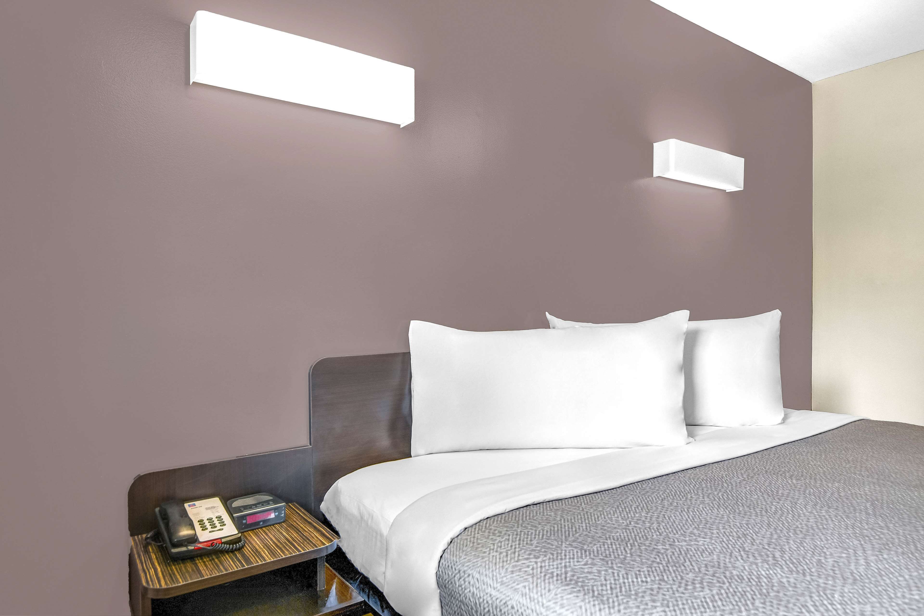 A hotel room with a bed, a bedside table with a phone and a small device, and two wall-mounted light fixtures.