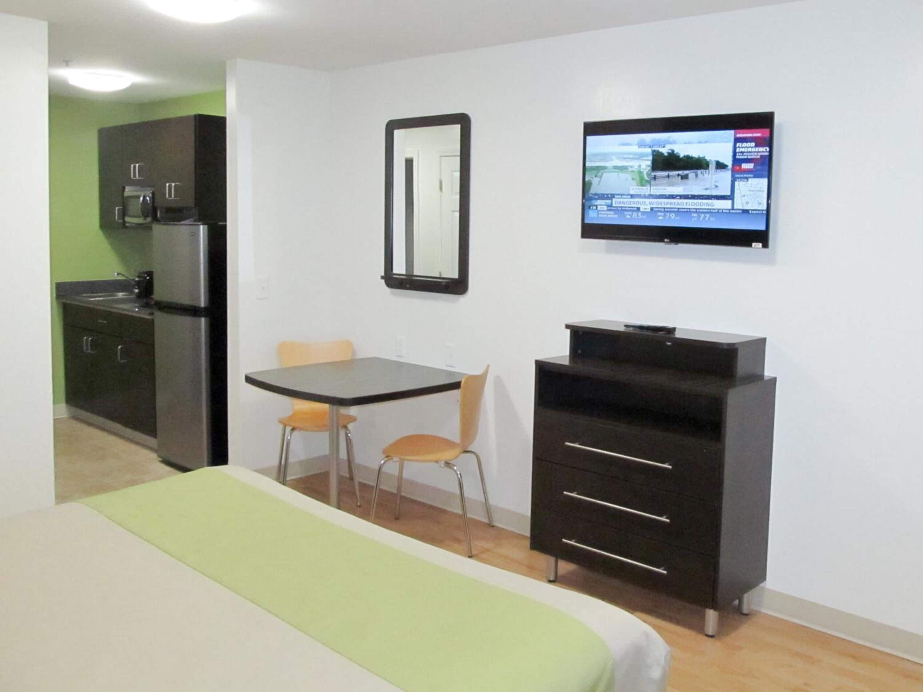 A compact hotel room with a bed, a small table with two chairs, a kitchenette, a TV mounted on the wall, and a mirror.