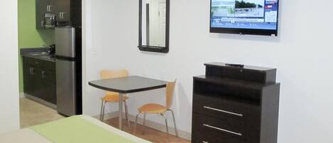 Quarto de hotel compacto com uma cama, uma mesinha com duas cadeiras, cozinha compacta, TV fixada na parede e um espelho.