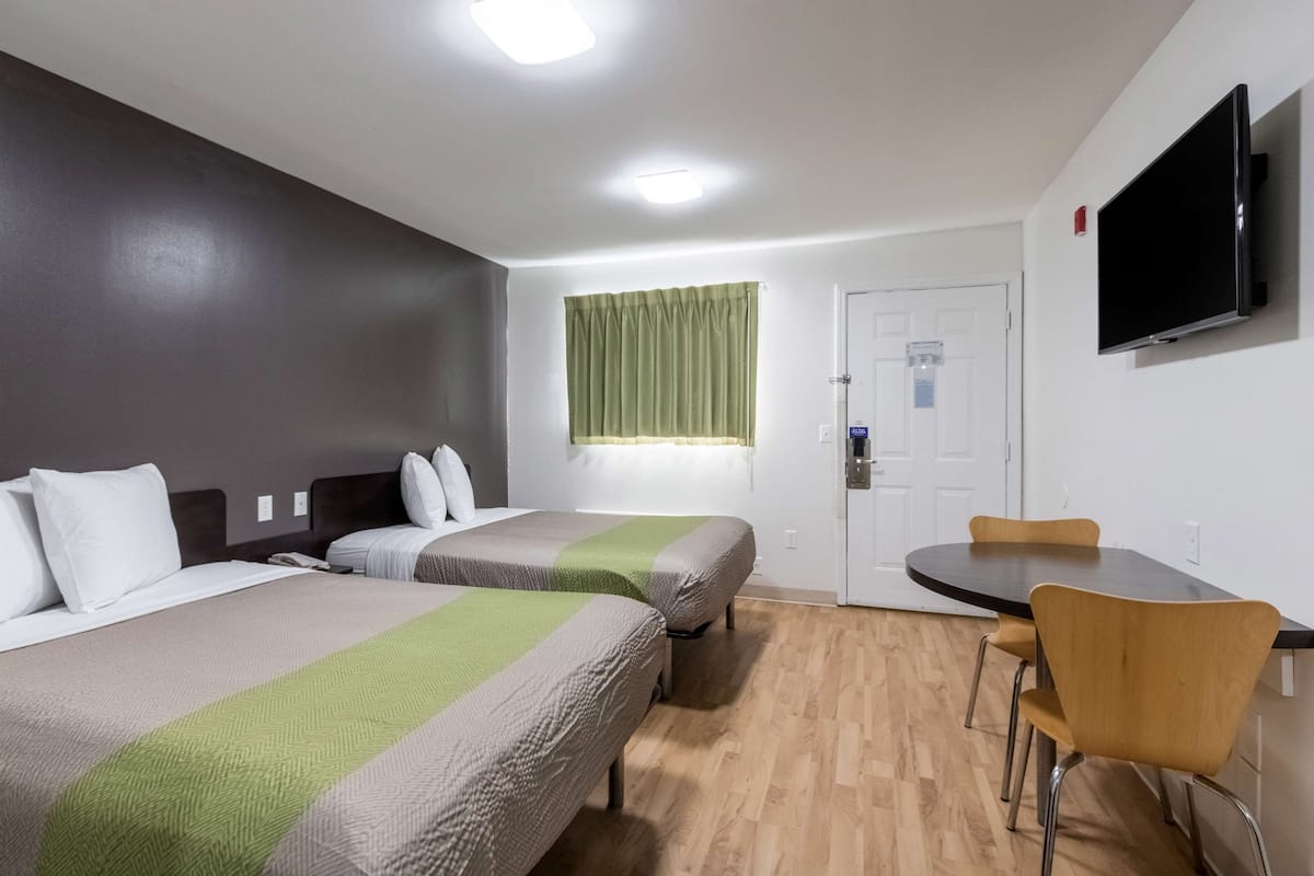 Habitación de hotel con dos camas, una mesa de comedor, sillas, televisor y ventana con cortinas.