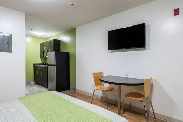 A hotel room with a bed, a small table with two chairs, a TV mounted on the wall, and a kitchenette in the background.