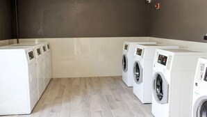 A laundry room with four white washing machines and a dryer, a grey wall, and a tiled floor. - Studio 6 Extended Stay - Savannah, GA (Savannah)