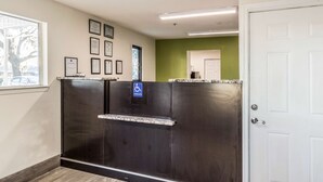 A reception area with a dark reception desk, framed certificates on the wall, and a wheelchair accessibility sign. - Studio 6 Extended Stay - Savannah, GA (Savannah)