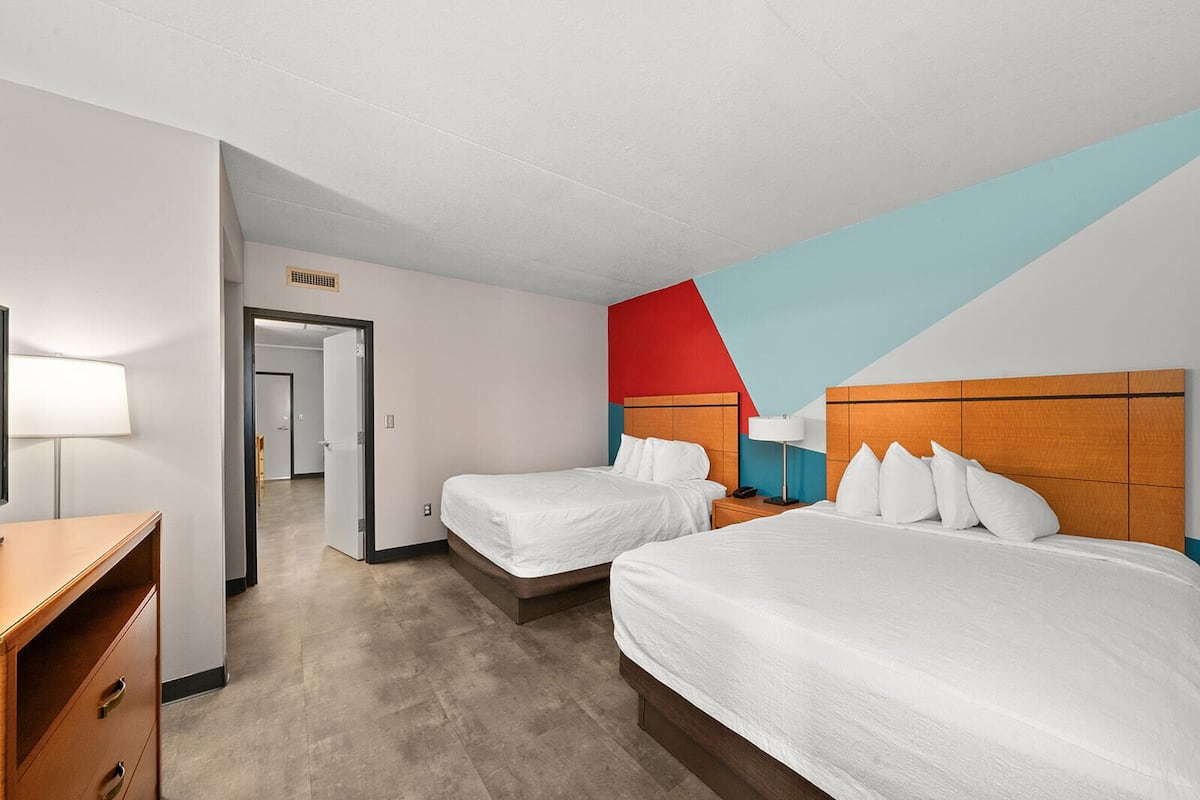 A hotel room with two beds, a desk, and a chair. There is a door leading to another room.