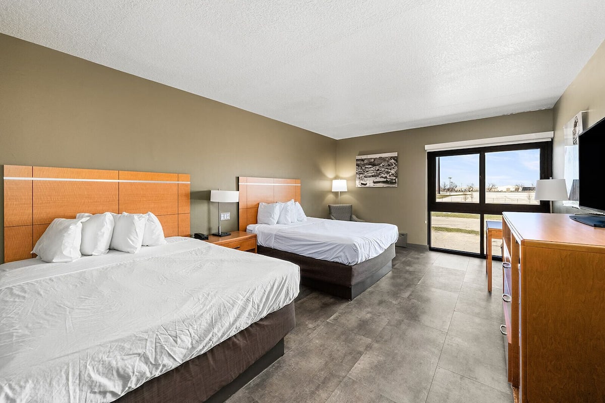 A hotel room with two beds, a desk, a chair, a TV, and a large window offering a view of the outdoors.