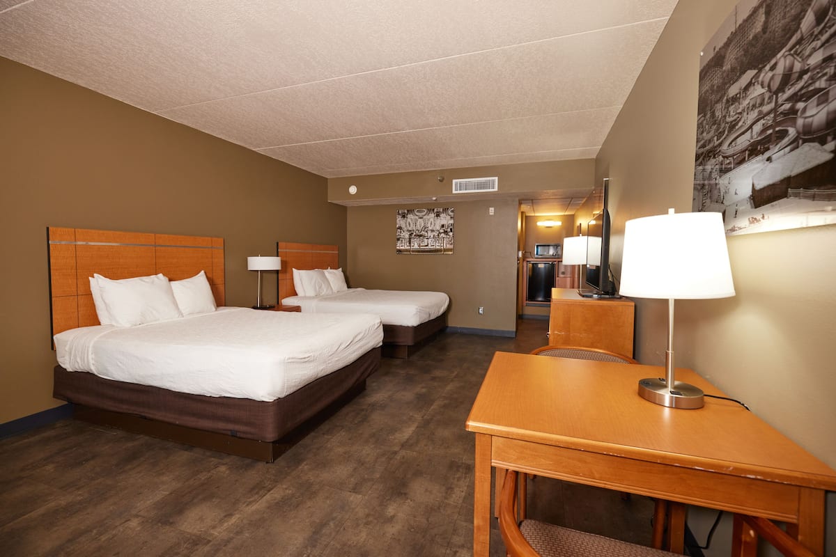 A hotel room with two beds, a desk, a chair, a lamp, and a wall-mounted TV.