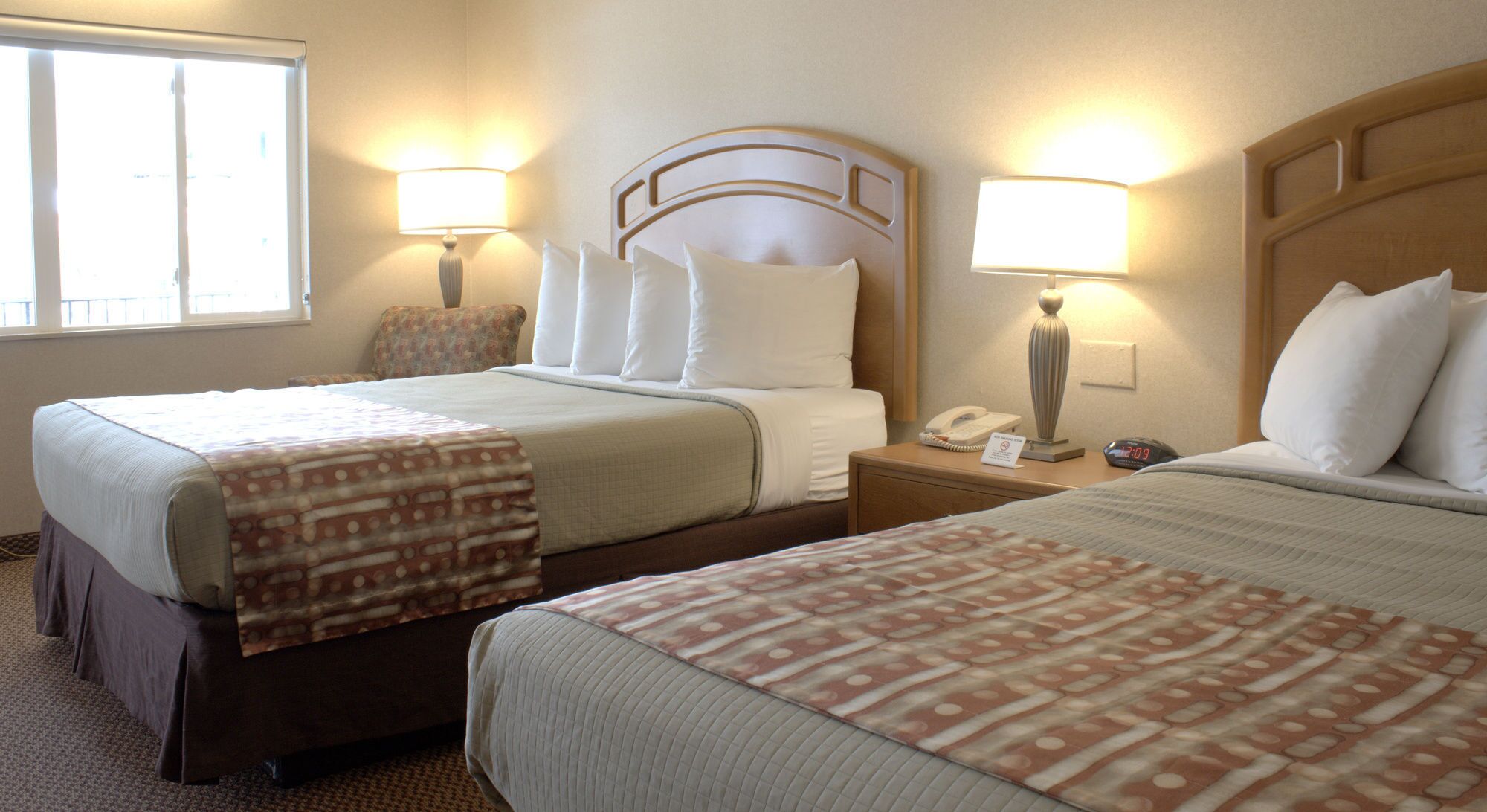 Habitación de hotel con dos camas, cada una con una colcha estampada, cabecero, lámparas de noche, mesita con teléfono y cartel, y ventana con persianas.