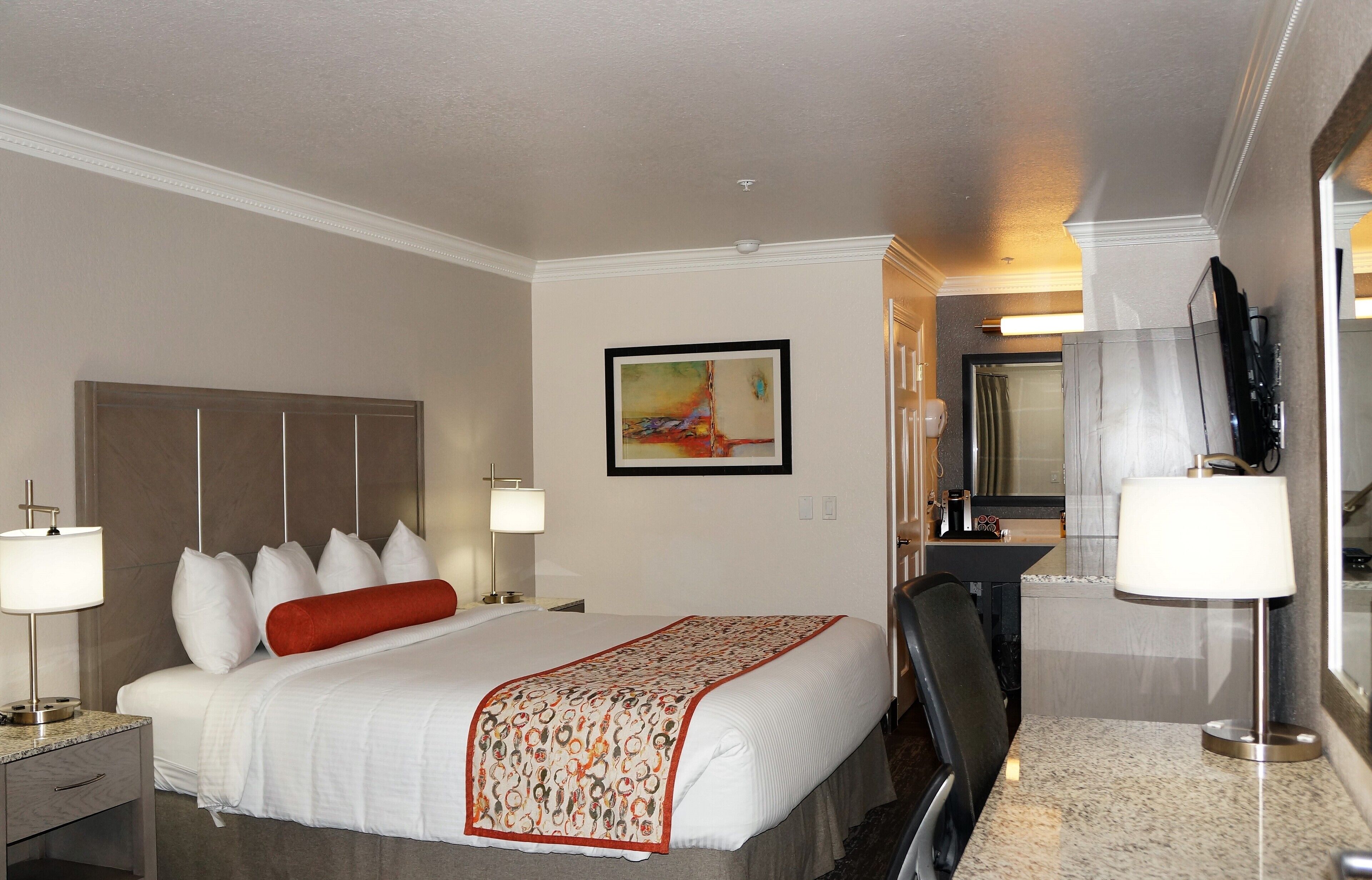 A hotel room with a large bed, two bedside tables with lamps, a desk with a chair, a television, and a painting on the wall.