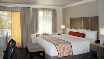 A hotel room with a large bed, a desk, and a chair. There is a window with curtains, a lamp, and a heater.