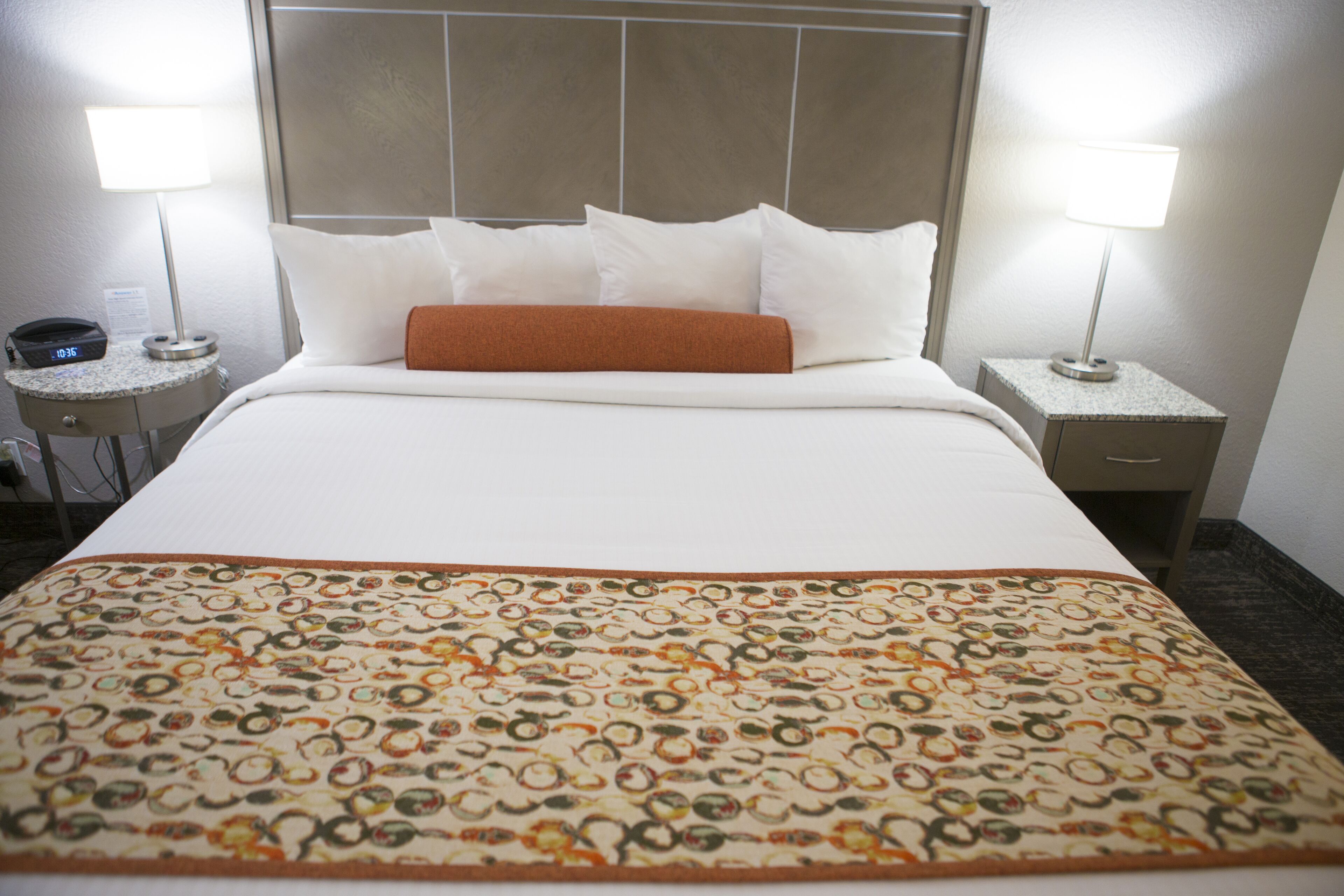 A hotel room with a neatly made bed, two bedside lamps, a clock, and a small table with a phone.