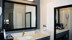 A modern bathroom with a large mirror, a coffee machine, and toiletries on the counter.