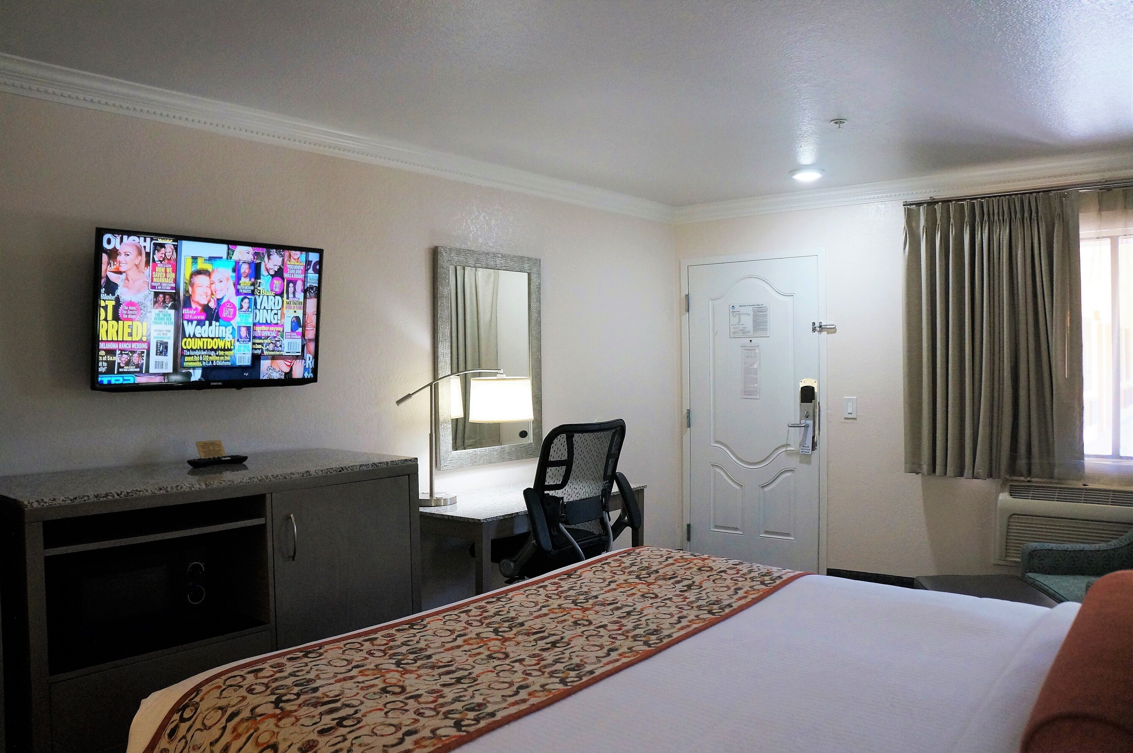A hotel room with a bed, a desk, a chair, a television mounted on the wall, and a door.