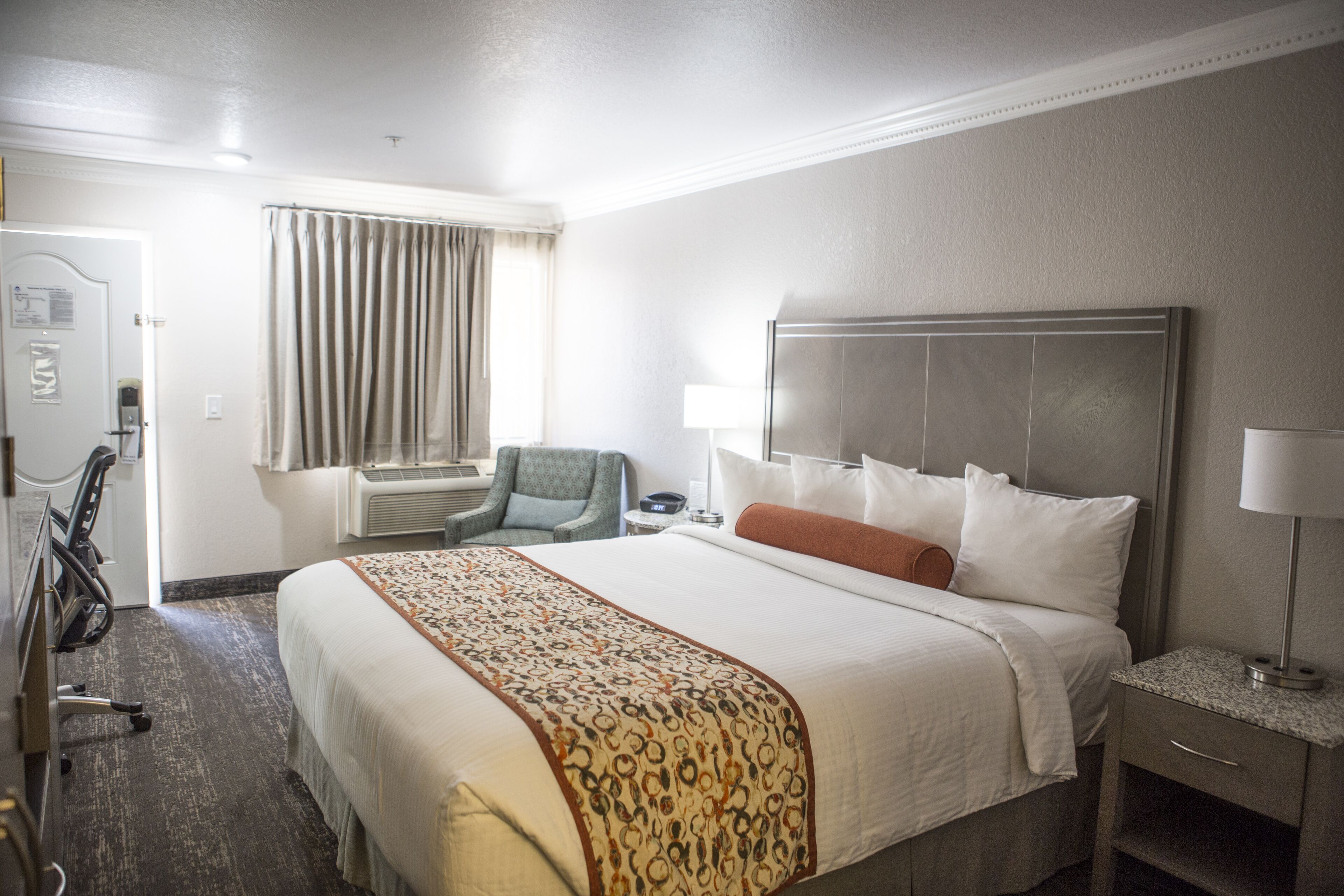 A hotel room with a large bed, a desk with a lamp, a chair, and a window with curtains.