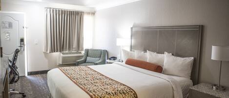 A hotel room with a large bed, a desk with a lamp, a chair, and a window with curtains.