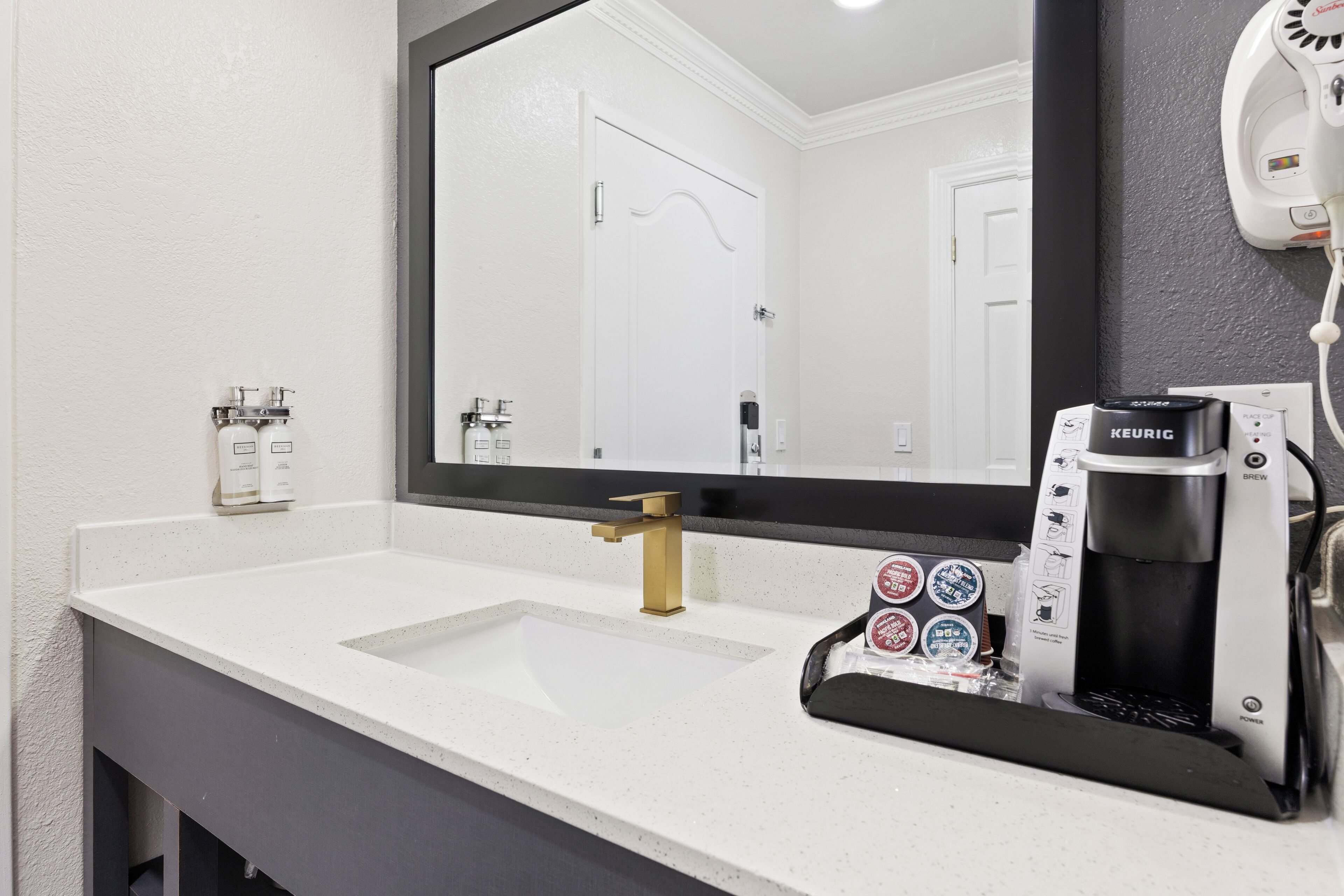 A bathroom with a white sink, a gold faucet, and a mirror. There is a coffee maker and a hairdryer on the wall.