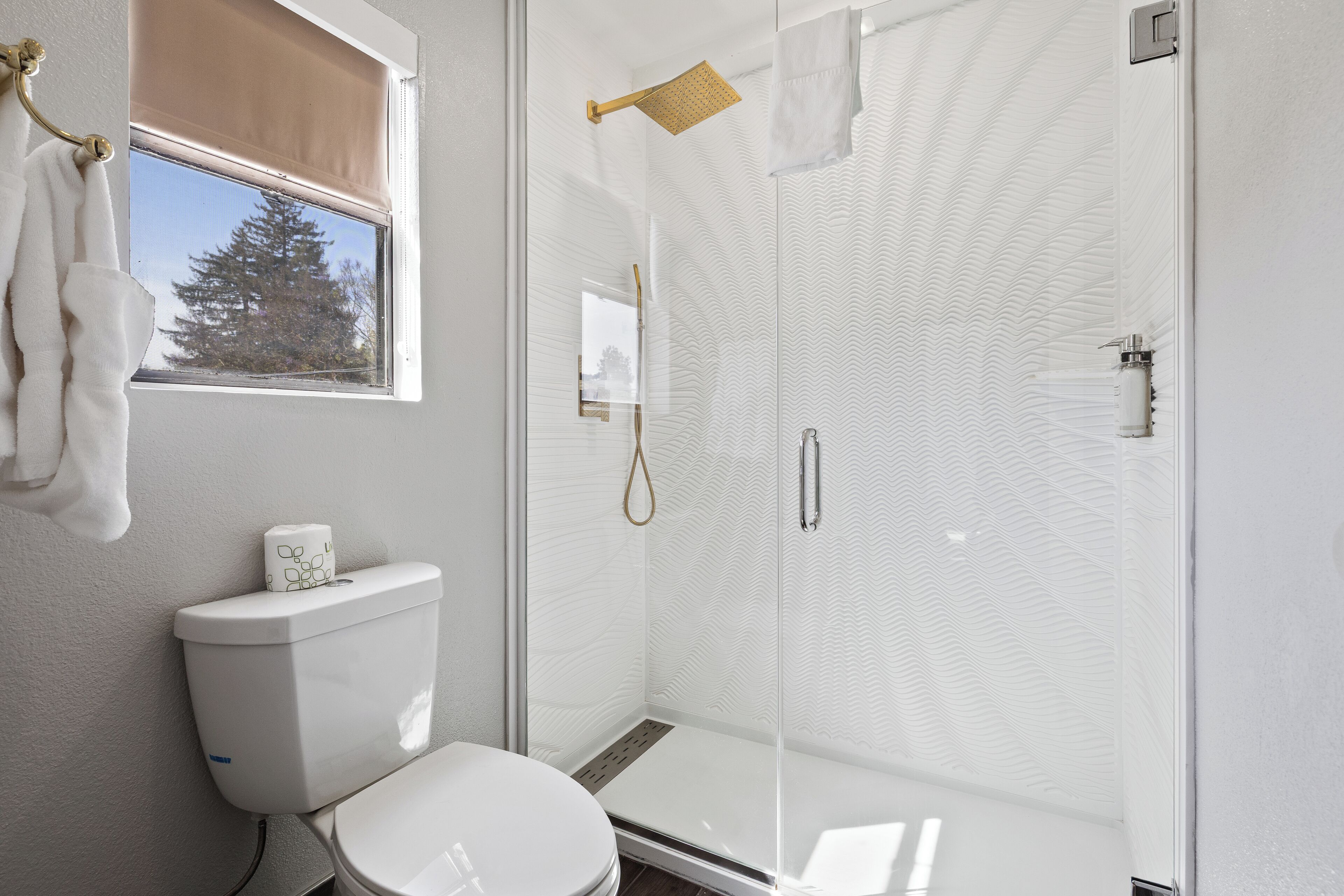 Un baño con inodoro, cabina de ducha de vidrio y ventana con vista a un árbol.