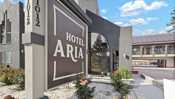 A modern hotel entrance with a large sign reading "HOTEL ARIA," a parking area, and a building with a solar panel roof.