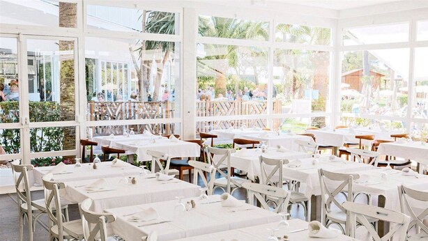 A restaurant with white tables and chairs, large windows, and a view of the outdoor seating area. - Hotel Globales Cala Blanca (Ciutadella de Menorca)
