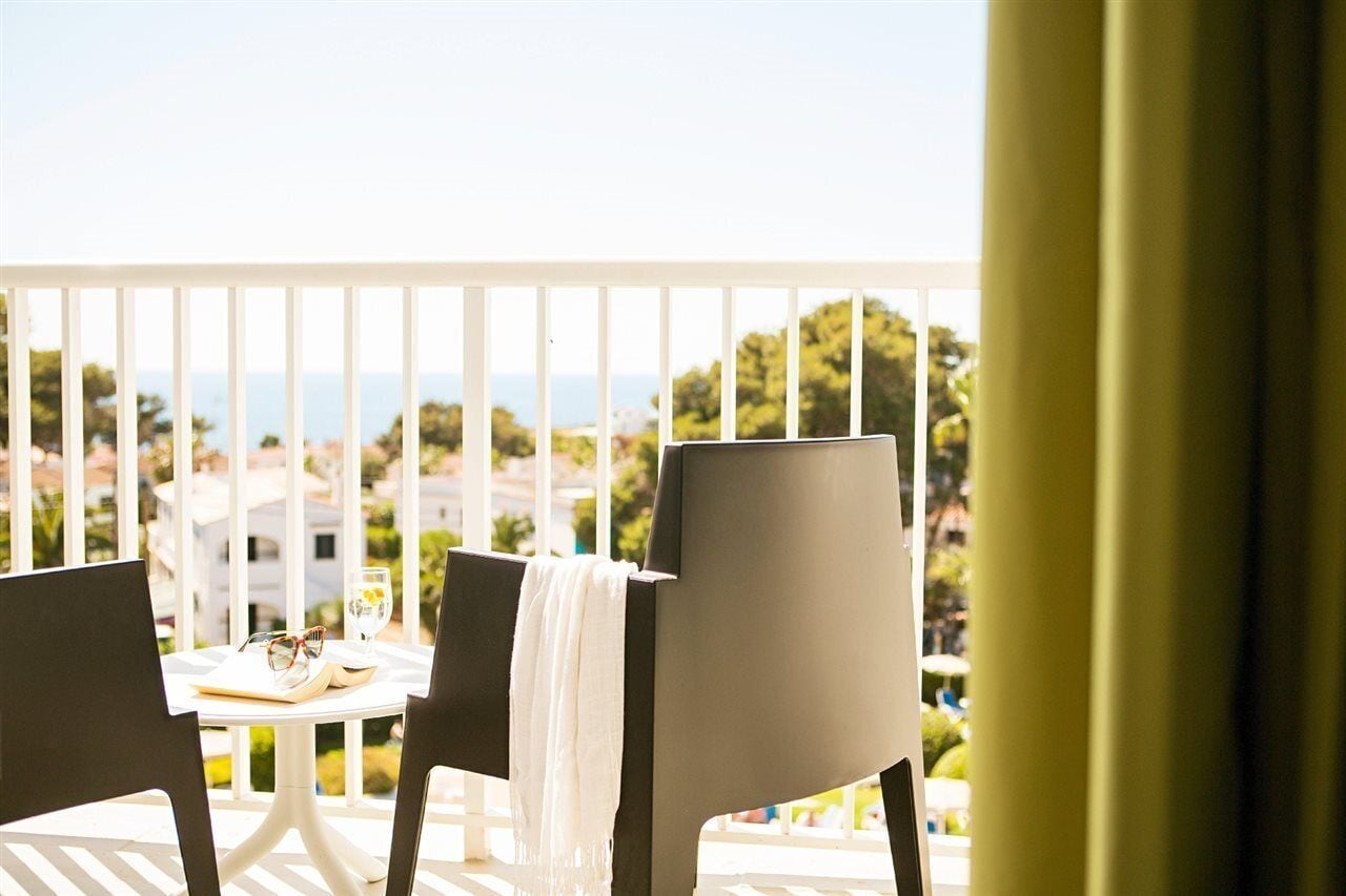 En balkon med to stole, et lille bord med en bog og glas, samt udsigt til havet og bygninger.