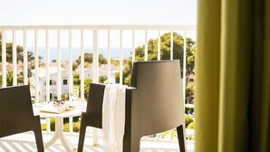 A balcony with two chairs, a small table with a book and glasses, and a view of the sea and buildings. - Hotel Globales Cala Blanca (Ciutadella de Menorca)