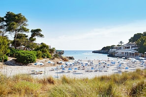 A beach with numerous sun loungers and umbrellas, a clear sky, and a rocky coastline. - Hotel Globales Cala Blanca (Ciutadella de Menorca)