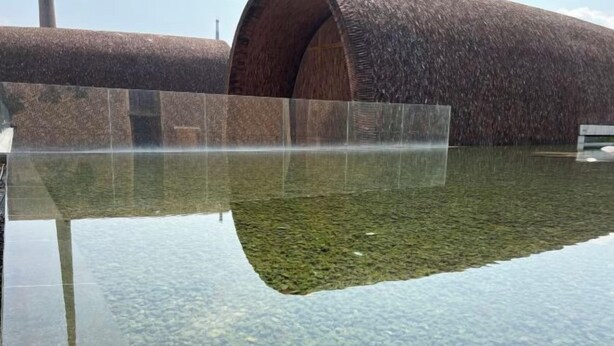 Un elemento acuático con agua cristalina, una barrera de vidrio, una zona ajardinada y una gran estructura cilíndrica.
