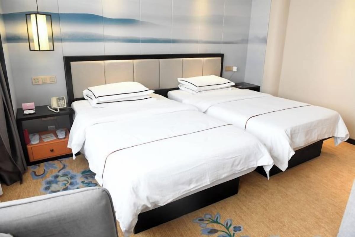 A hotel room with two beds, a nightstand, a lamp, and a view of the sky.