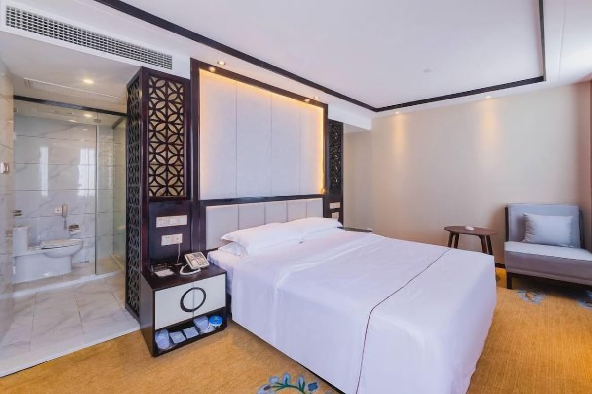 A hotel room with a bed, a bedside table, a nightstand, a chair, a small table, a mirror, a wardrobe, and a shower area.
