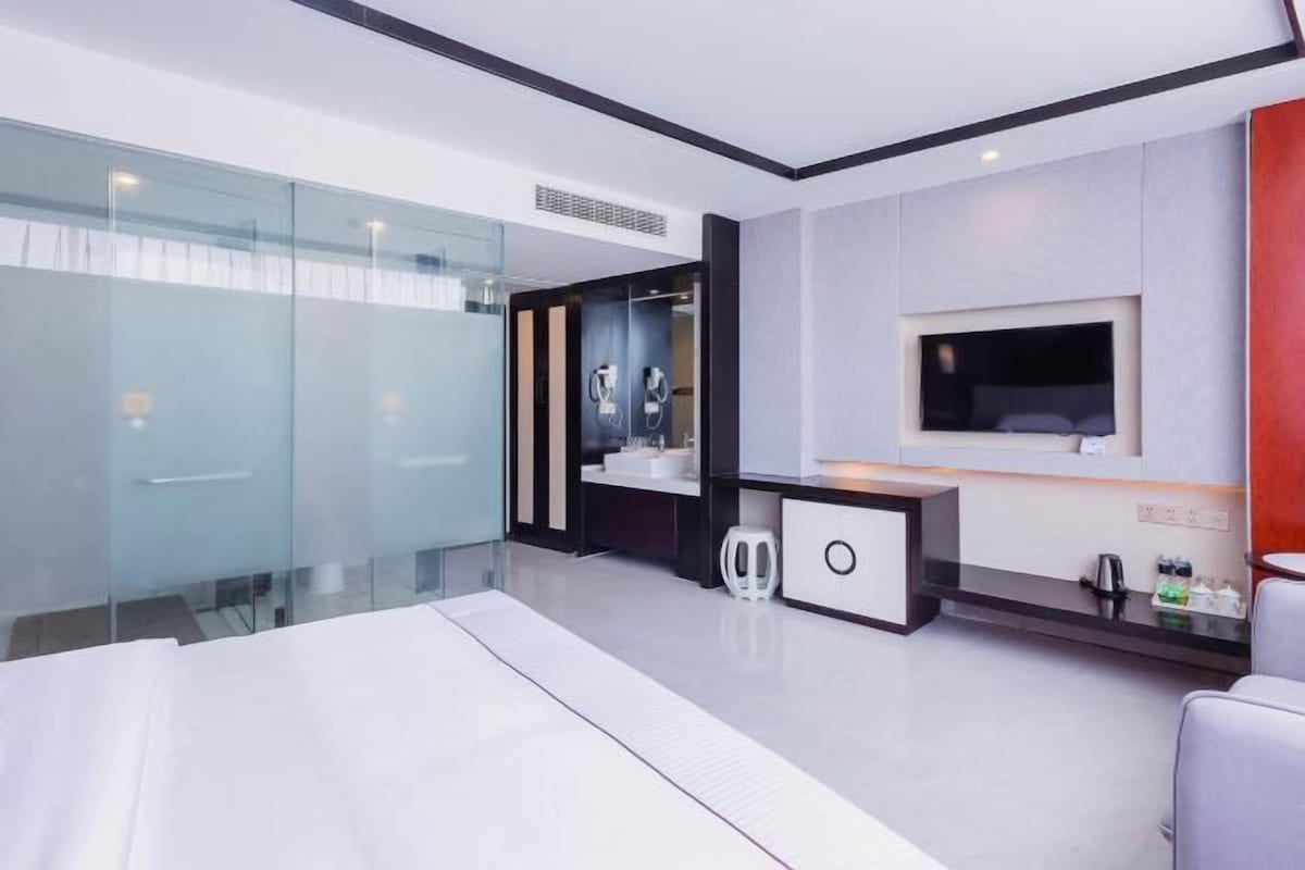 A modern hotel room with a bed, a TV, a bathroom with a glass shower, and a small seating area.
