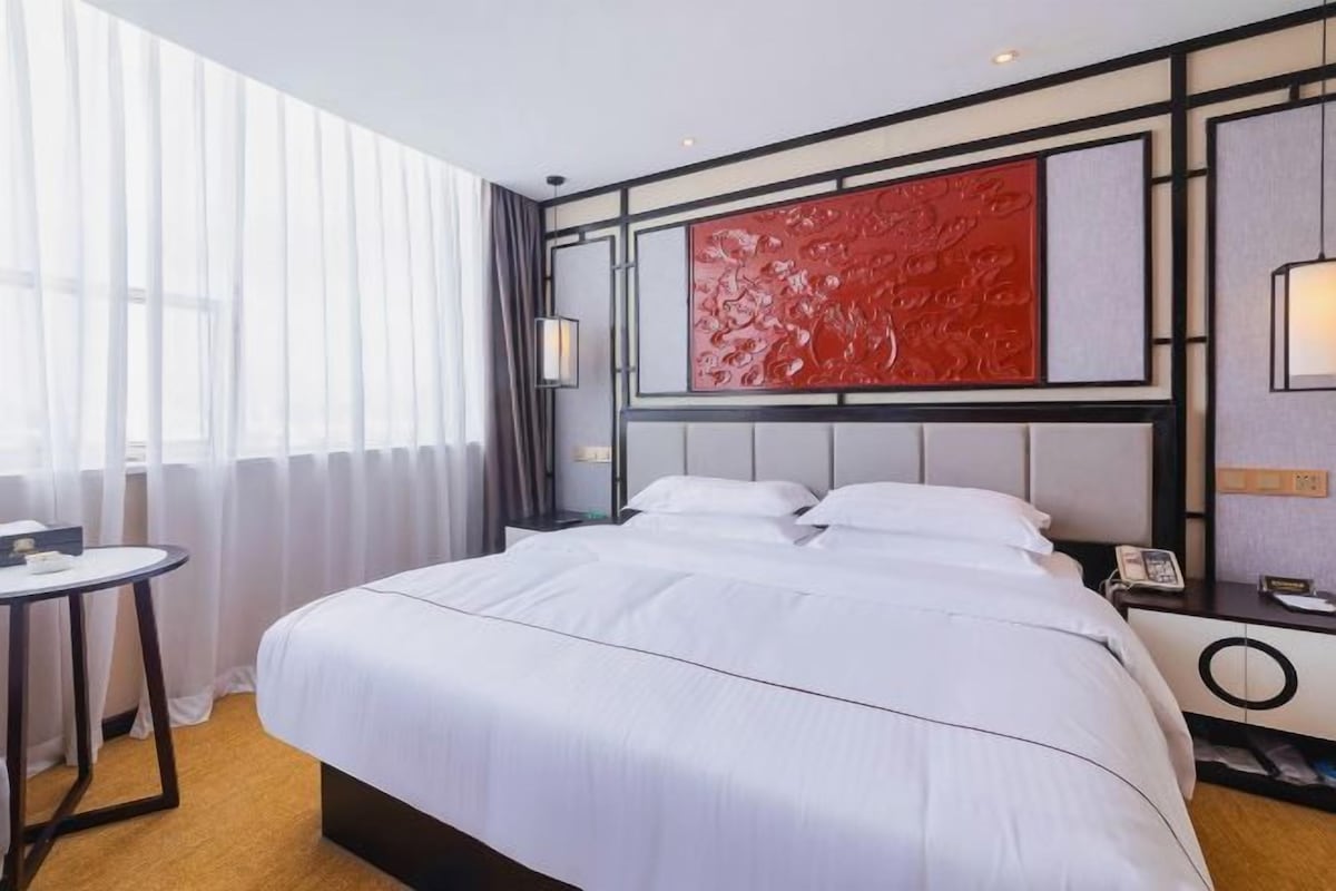 A hotel room with a large bed, a bedside table, a lamp, and a window with sheer curtains.