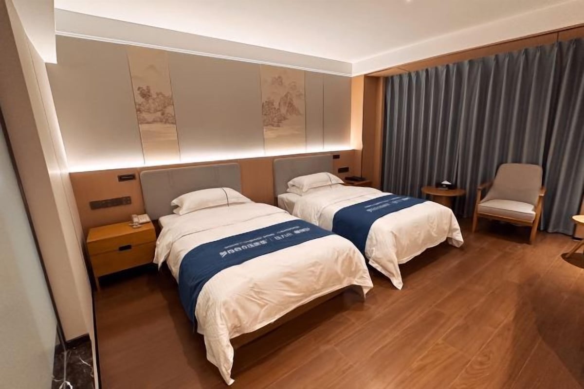 A hotel room with two beds, a nightstand, a chair, and a large window with curtains.