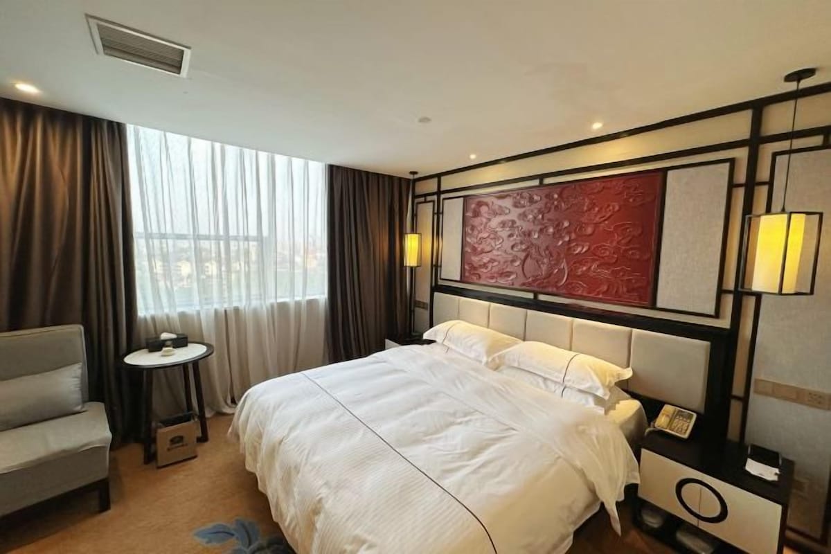 A hotel room with a large bed, a bedside table, a chair, and a view of the outdoors.