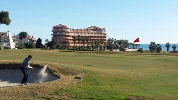 A golf course with a sand bunker, a red flag, and a building with balconies in the background.
