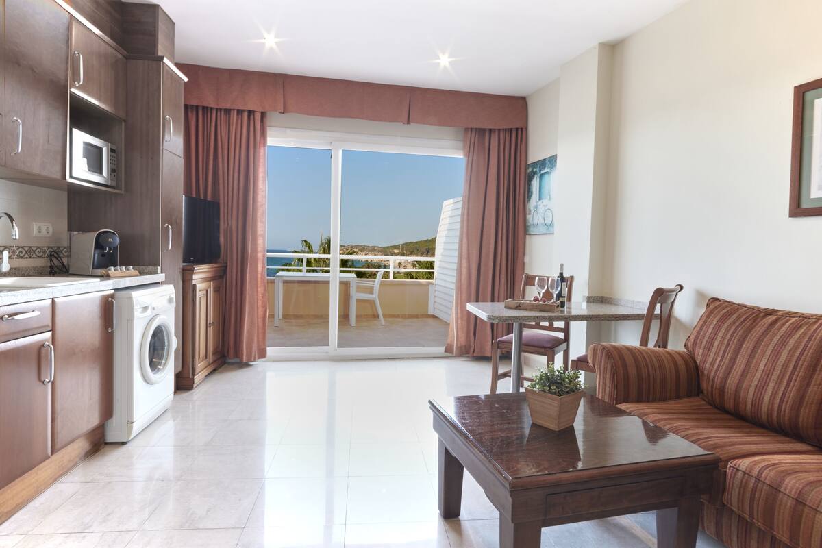 A modern kitchen with a washing machine, a dining area with a table and chairs, and a balcony with a view of the sea.