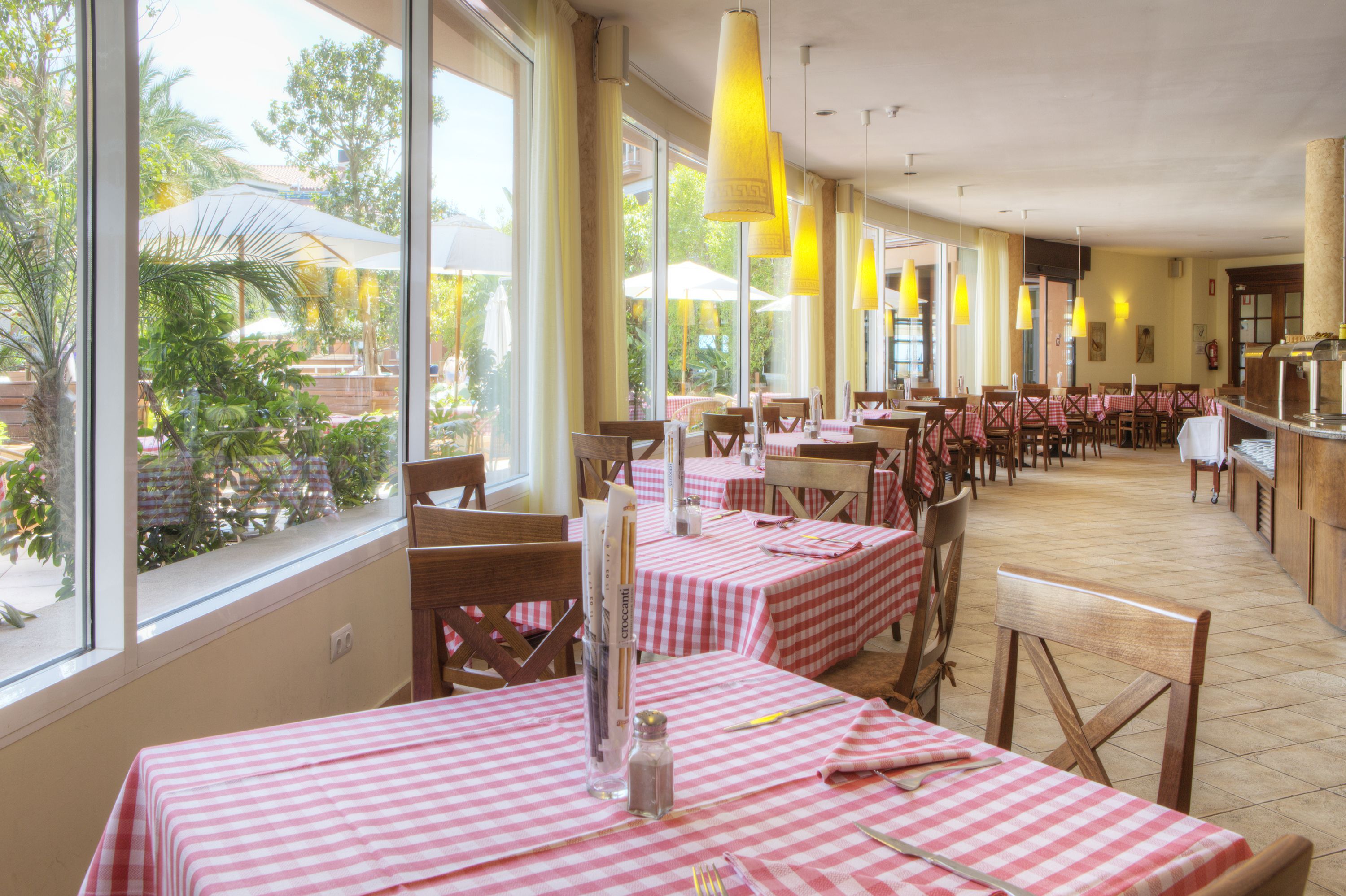 Un restaurante con mesas a cuadros rojos y blancos, ventanas grandes y lámparas colgantes amarillas.
