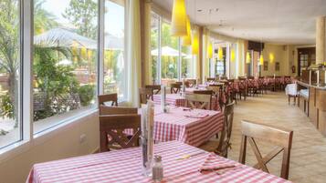 Un restaurante con mesas a cuadros rojos y blancos, ventanas grandes y lámparas colgantes amarillas.