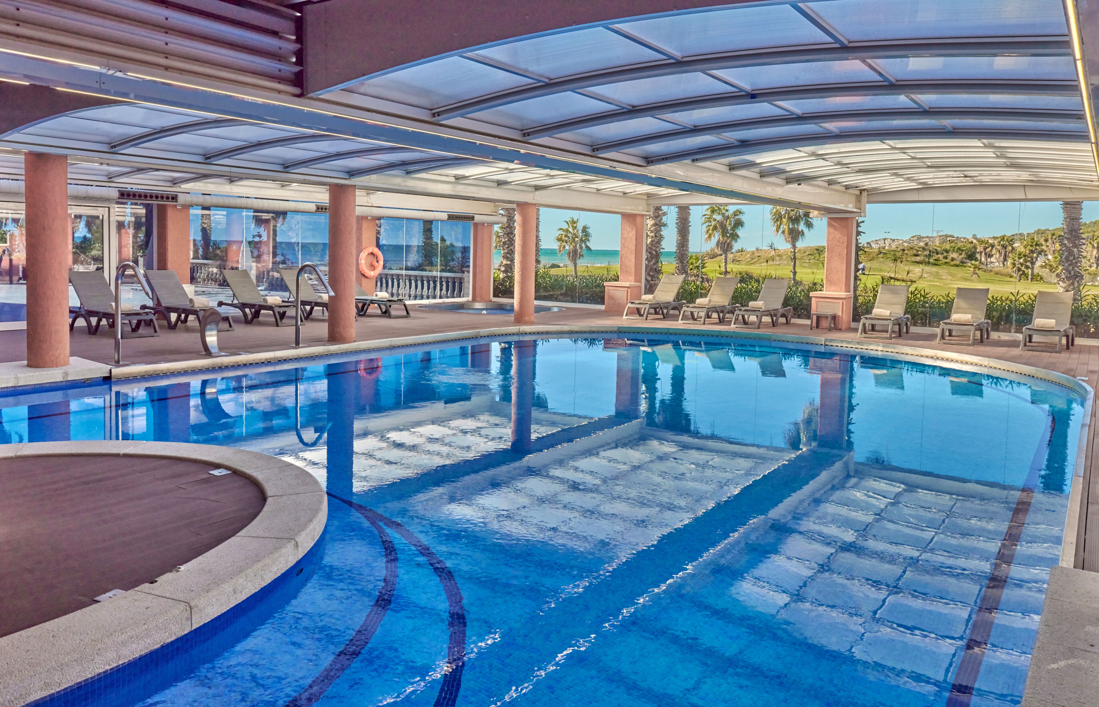 Uma grande piscina interna com  água azul cristalina, rodeada por espreguiçadeiras e um teto de vidro.