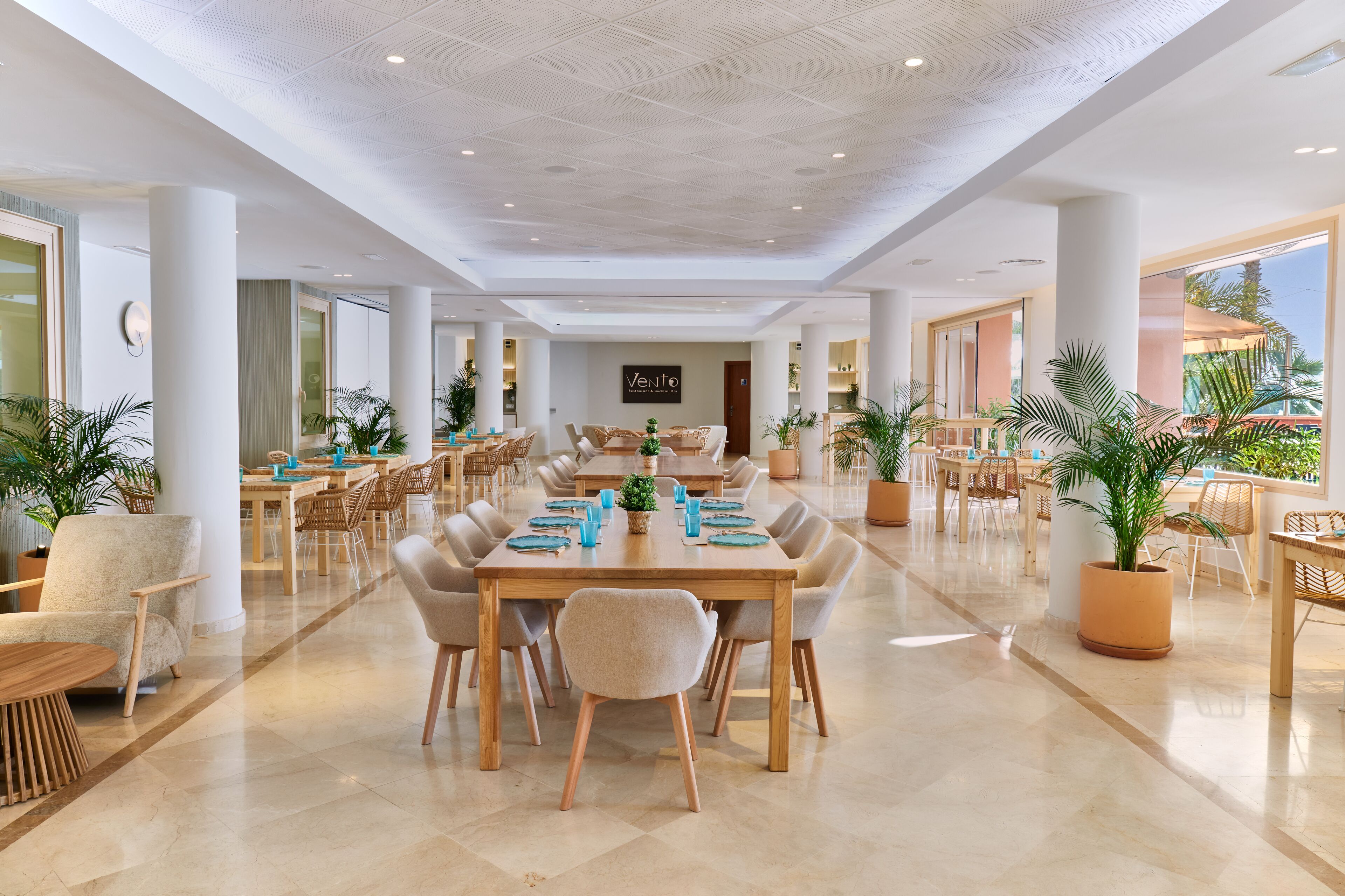 A spacious hotel lobby with multiple dining areas, large windows, and potted plants.