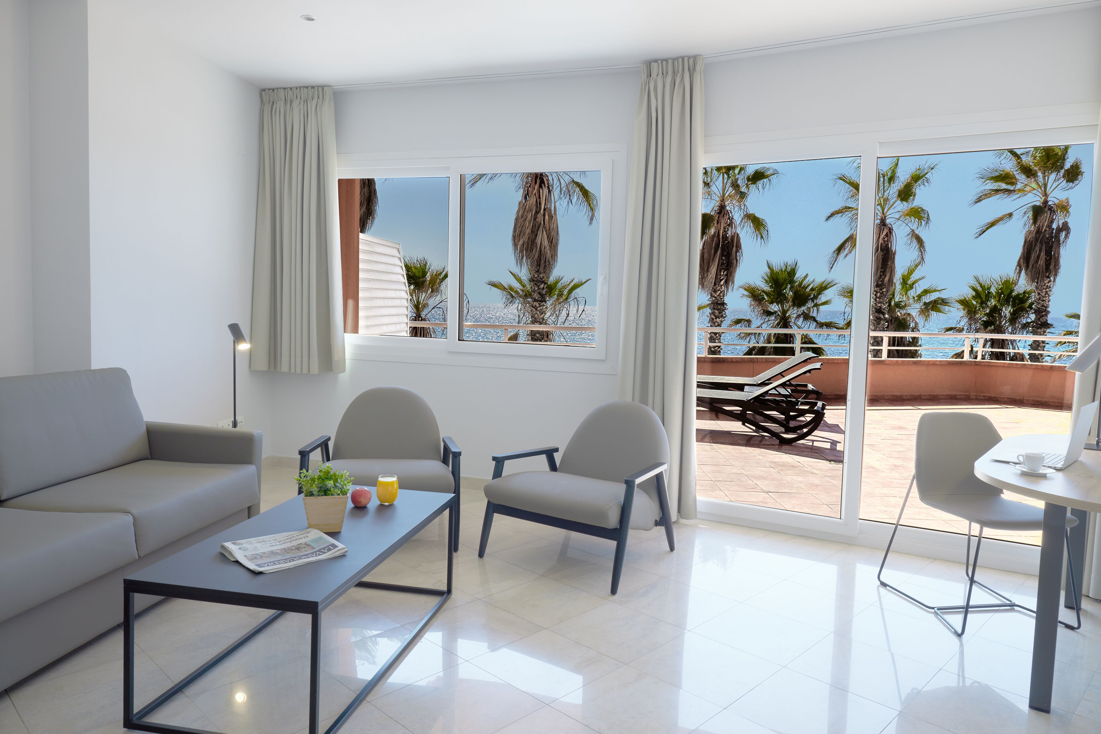 Una sala de estar moderna con un sofá, dos sillones, una mesa de centro y una mesa de comedor. Amplias ventanas ofrecen vistas a palmeras y un área de patio.