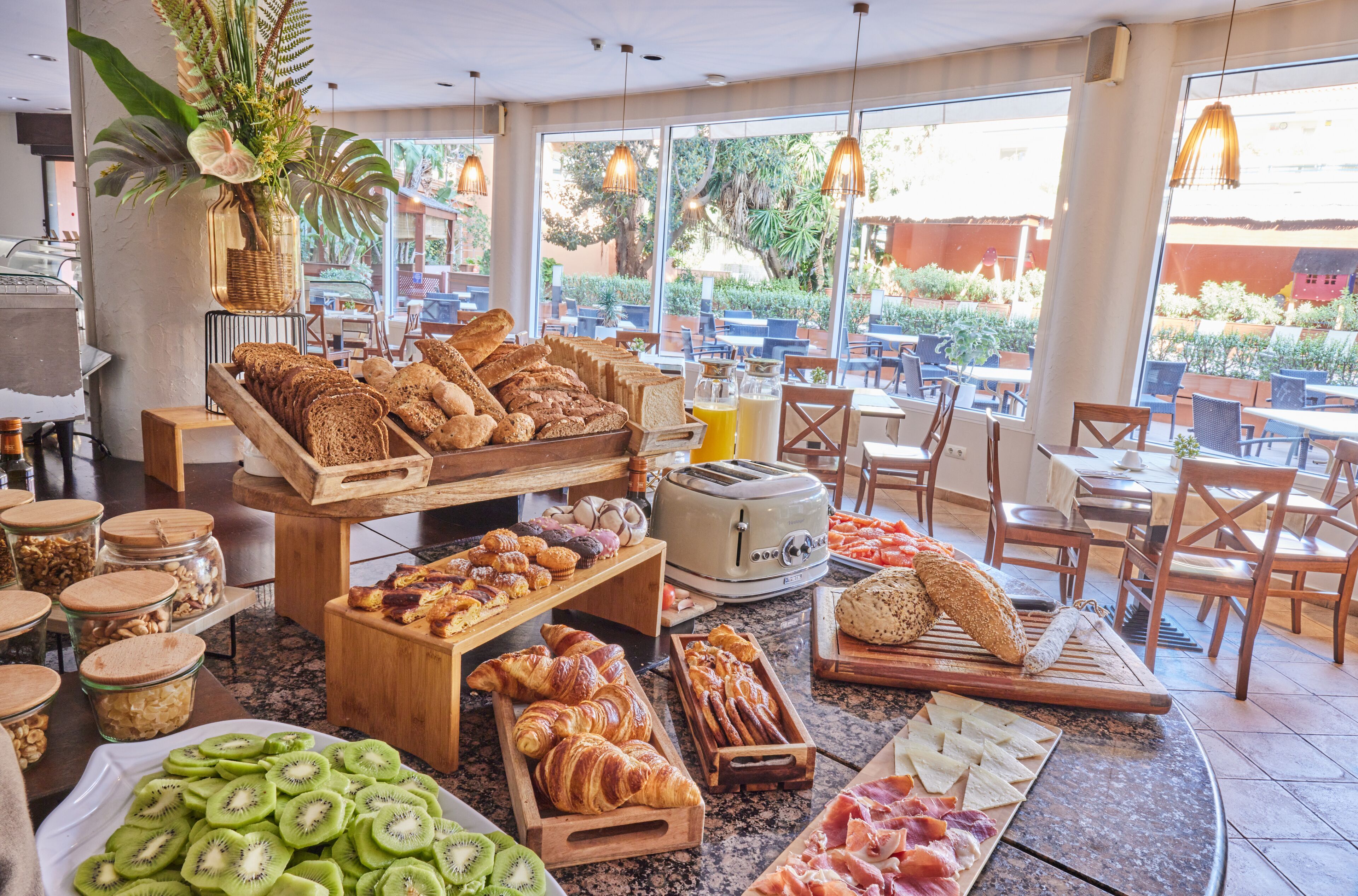 Un buffet de petit-déjeuner proposant une variété d’aliments, notamment des viennoiseries, du pain et des fruits, dans un espace intérieur bien éclairé et doté de grandes fenêtres.