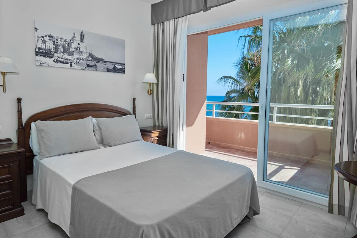 A hotel room with a bed, a nightstand, a lamp, a painting, and a balcony with a view of the sea and palm trees.