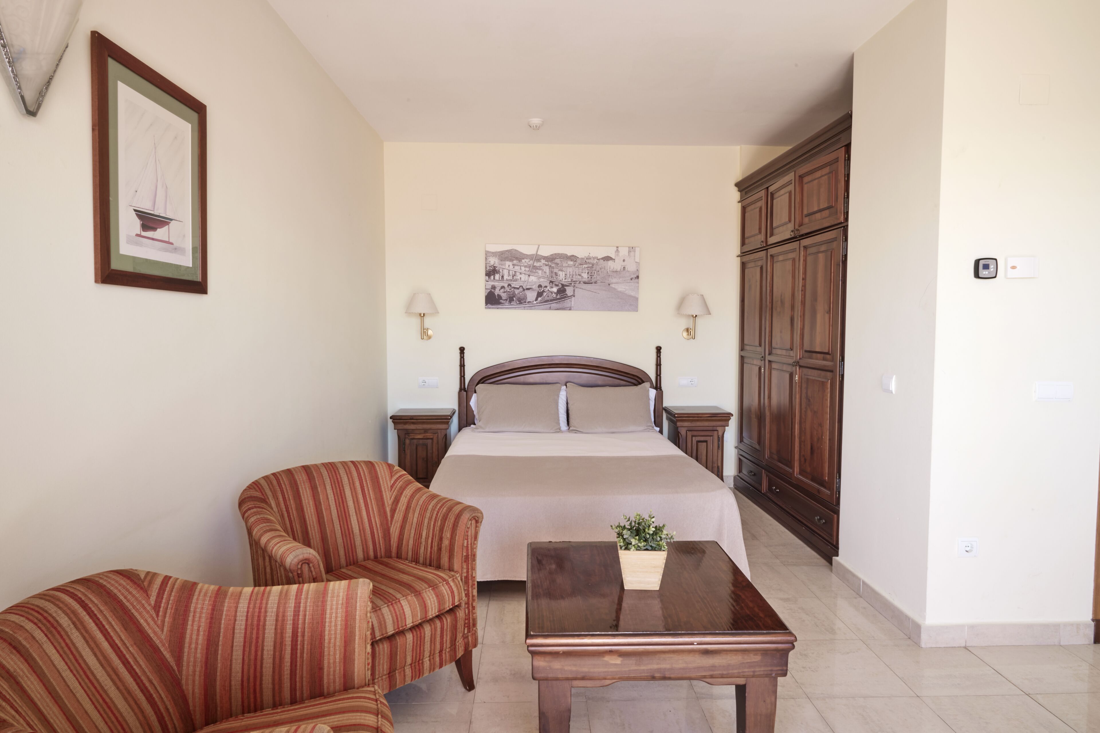 Un dormitorio con una cama, dos mesitas de noche, un armario de madera, un sillón a rayas, una mesa de madera y dos lámparas fijadas a la pared.